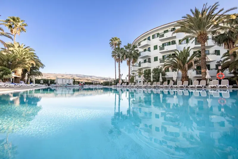 Une piscine turquoise reflète un bâtiment blanc et des palmiers sous un ciel bleu.