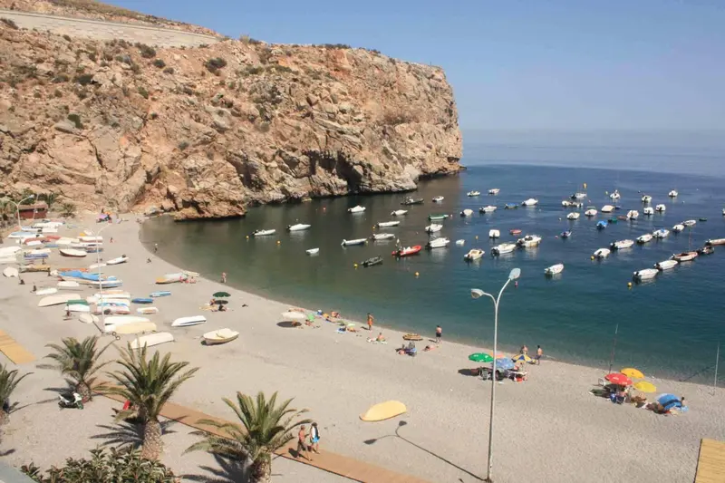 Une plage de sable avec des transats et des parasols, bordée par une falaise et une mer bleue.