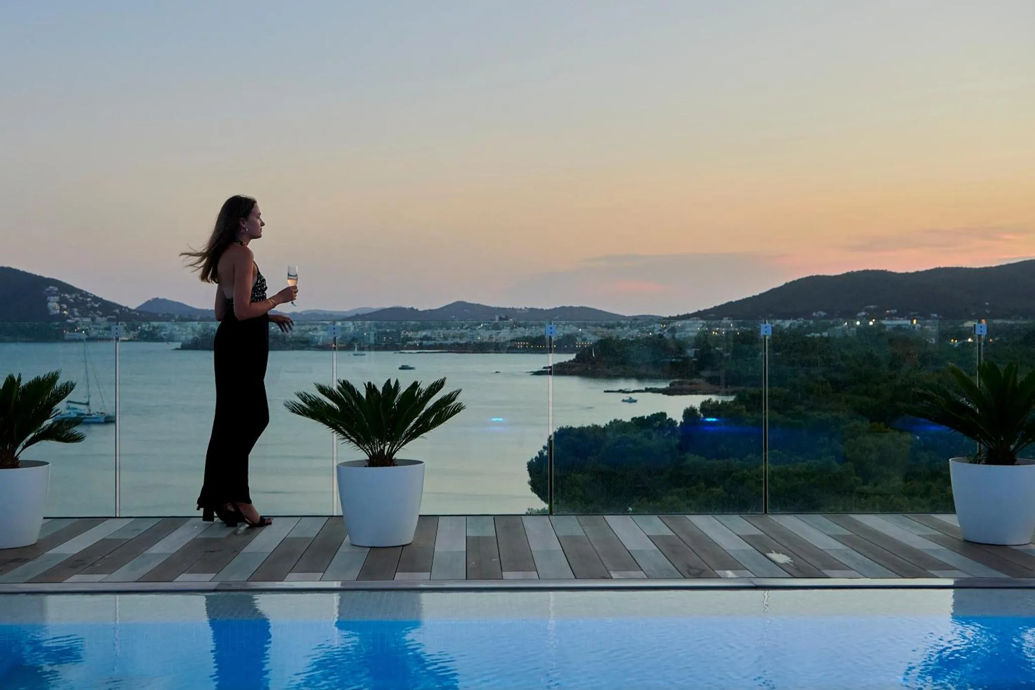Une femme admire le coucher de soleil sur la mer depuis une terrasse avec piscine.