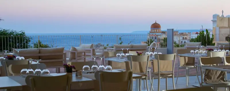 Terrasse aménagée avec tables et canapés face à la mer au coucher du soleil.