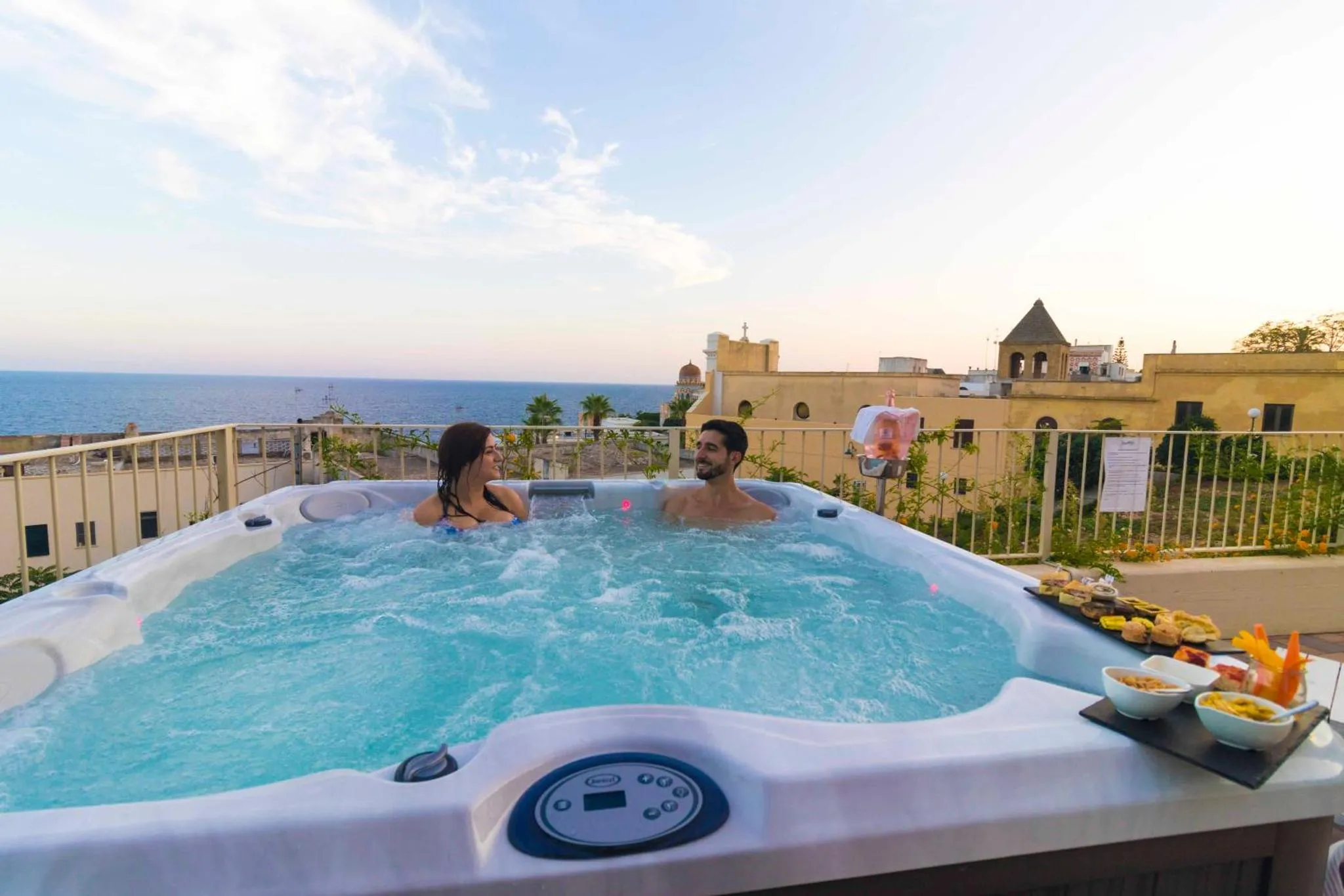 Un couple se détend dans un jacuzzi sur une terrasse avec vue sur la mer et un paysage urbain.