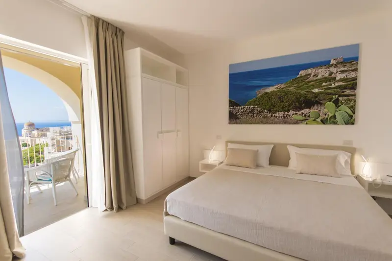 Chambre lumineuse avec un grand lit et une vue sur la mer depuis un balcon.