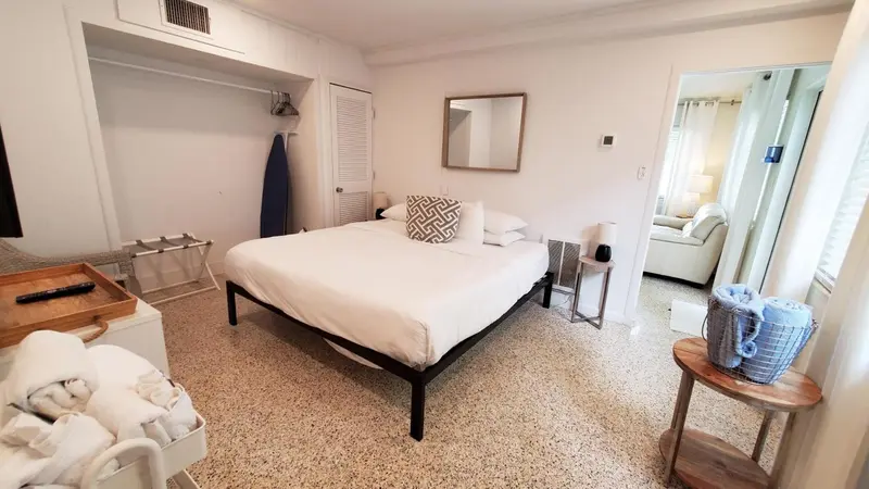 Une chambre lumineuse avec un lit fait et un espace salon visible au fond.