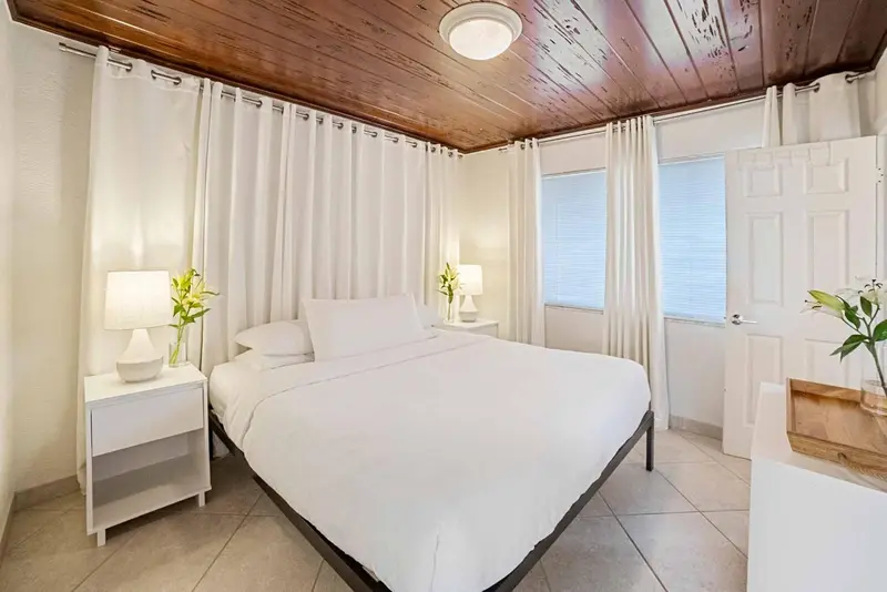 Une chambre lumineuse avec un lit blanc et des rideaux clairs.