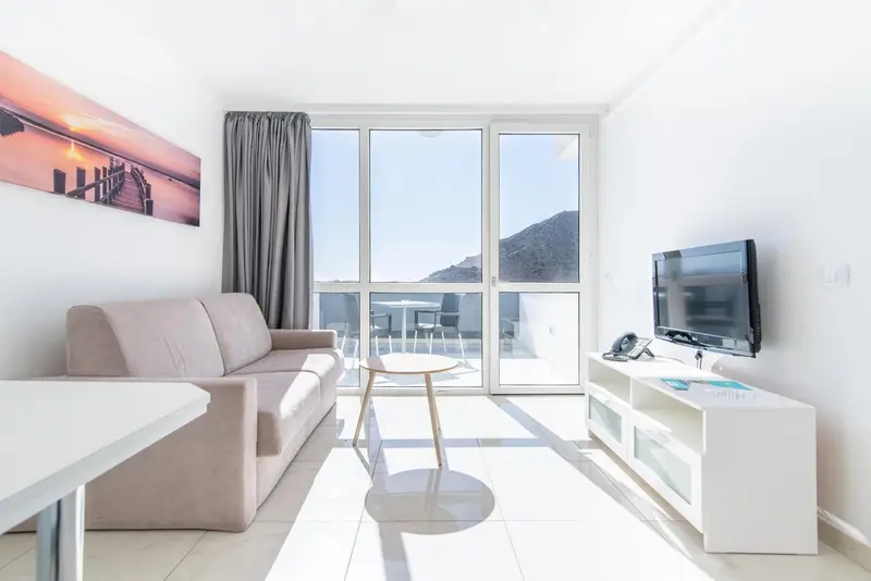 Un salon lumineux avec canapé beige et télévision murale donne sur une vue dégagée.