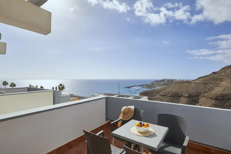 Une terrasse avec une table et des chaises offre une vue sur la mer et les collines.