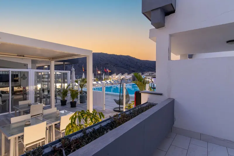Terrasse avec tables et chaises donnant sur une piscine et un paysage montagneux au coucher du soleil.