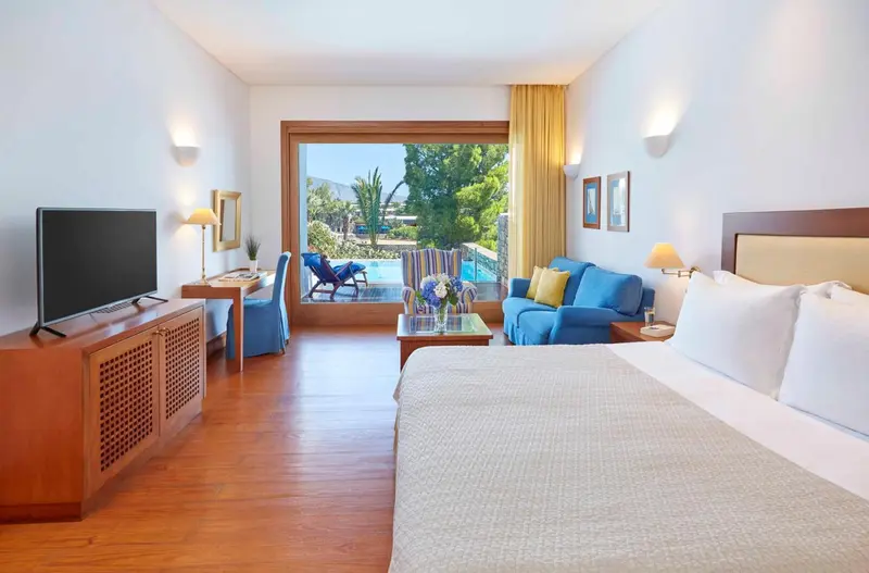 Chambre lumineuse avec un grand lit et un canapé bleu, donnant sur une piscine.