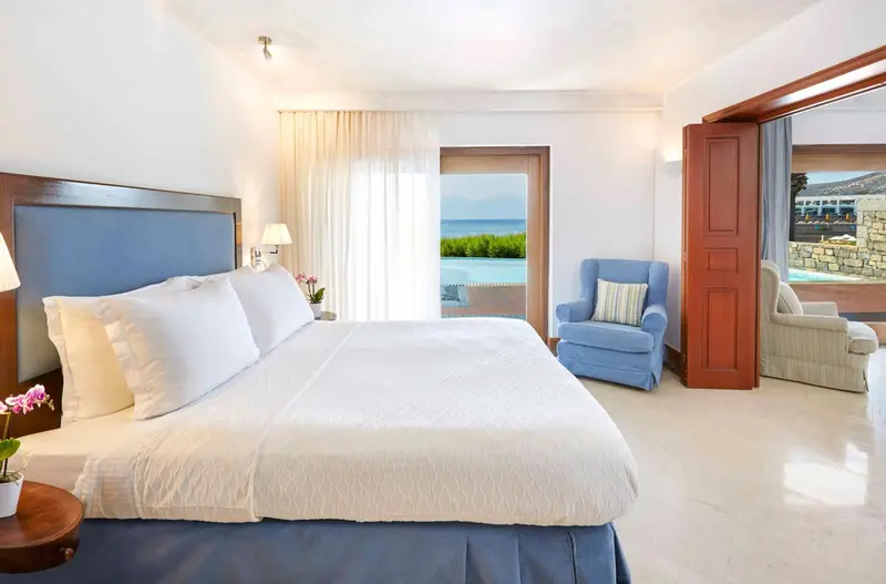 Chambre lumineuse avec un grand lit et un fauteuil près d'une baie vitrée.