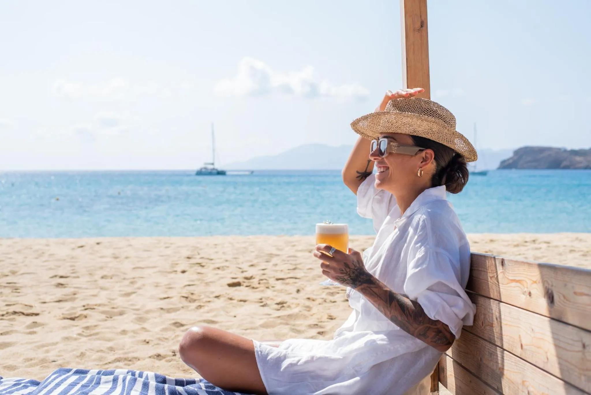 Une personne souriante se détend sur une plage ensoleillée avec une boisson.