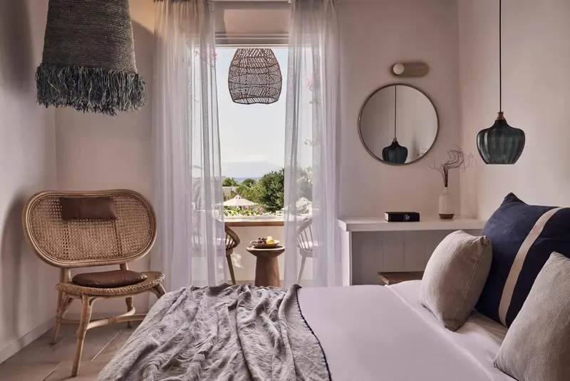 Une chambre lumineuse avec un lit et un fauteuil en rotin donnant sur une vue extérieure.