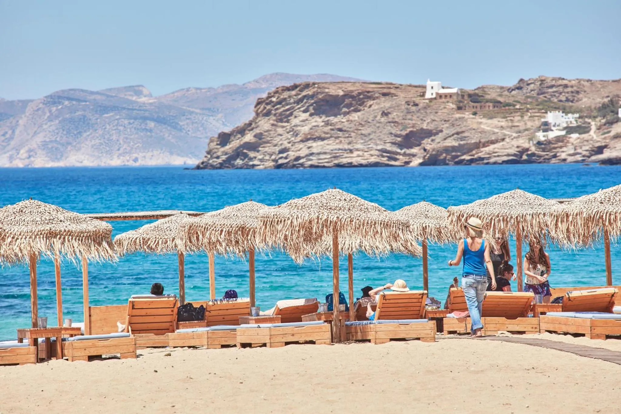 Transats et parasols sur une plage de sable fin avec vue sur la mer et des collines arides.