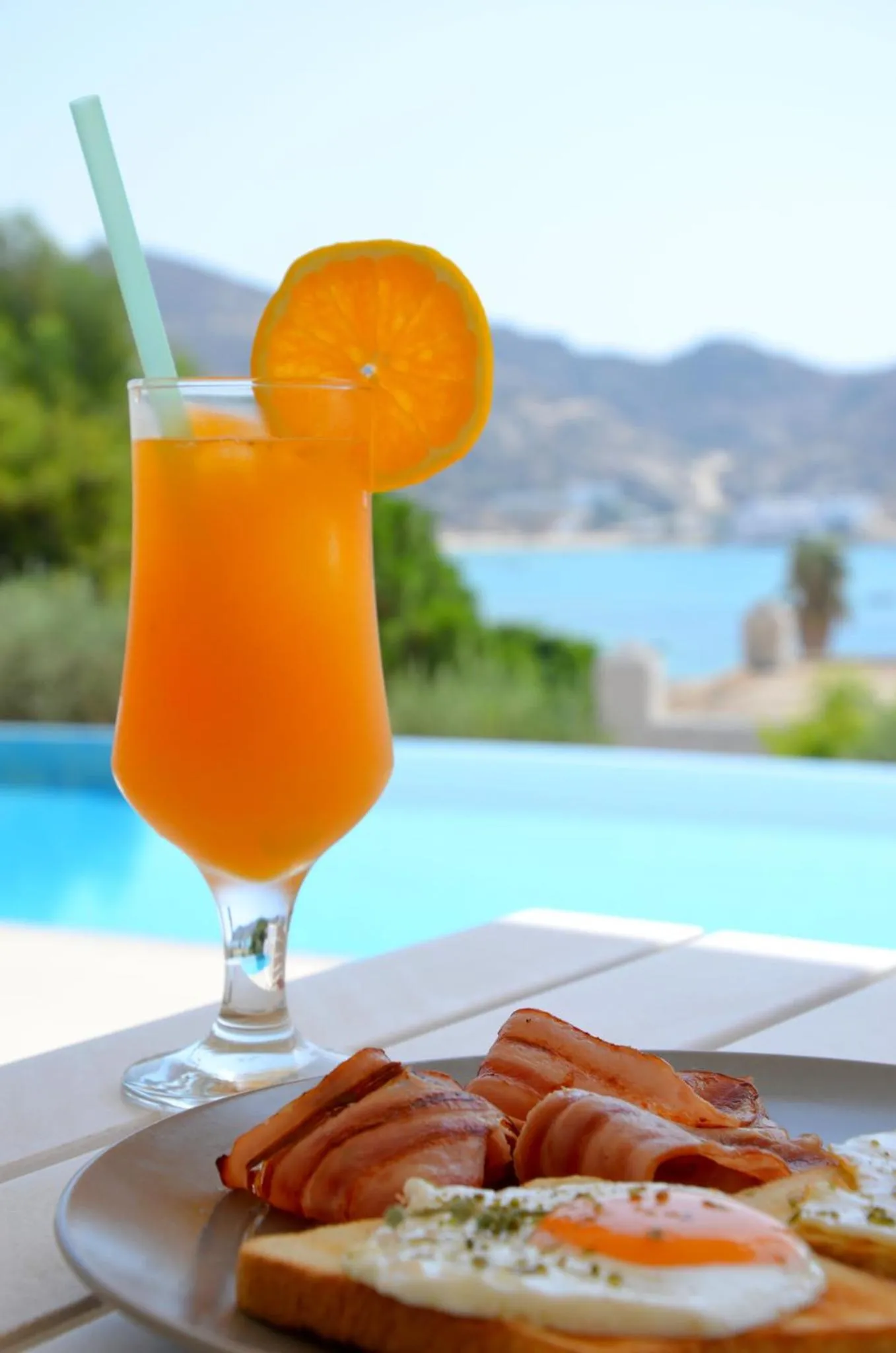 Un verre de jus d'orange et une assiette de nourriture sont posés au bord d'une piscine.