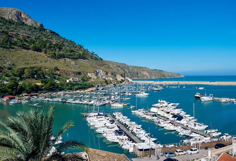 Vue aérienne d'un port de plaisance ensoleillé bordé de montagnes verdoyantes et de la mer turquoise.
