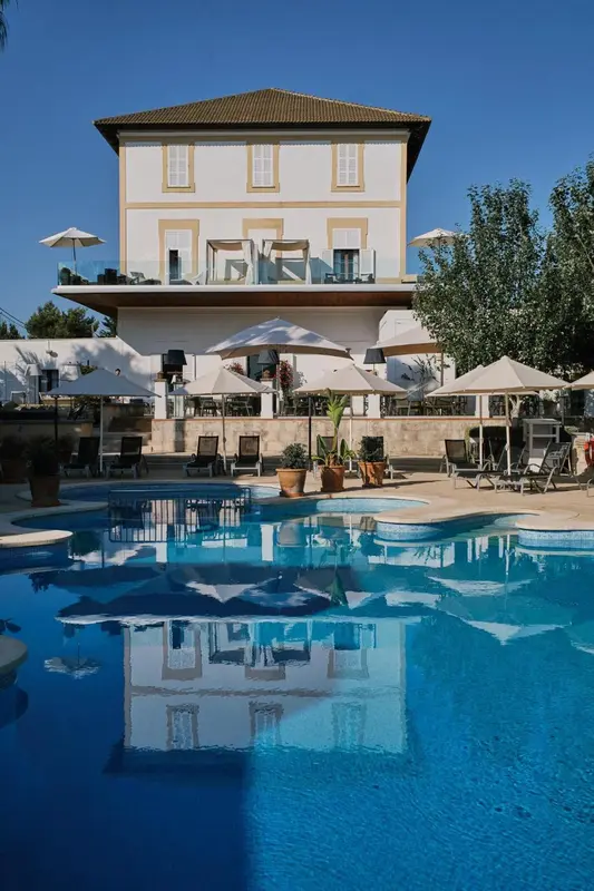 Bâtiment blanc à deux étages surplombant une piscine et une terrasse ensoleillée.