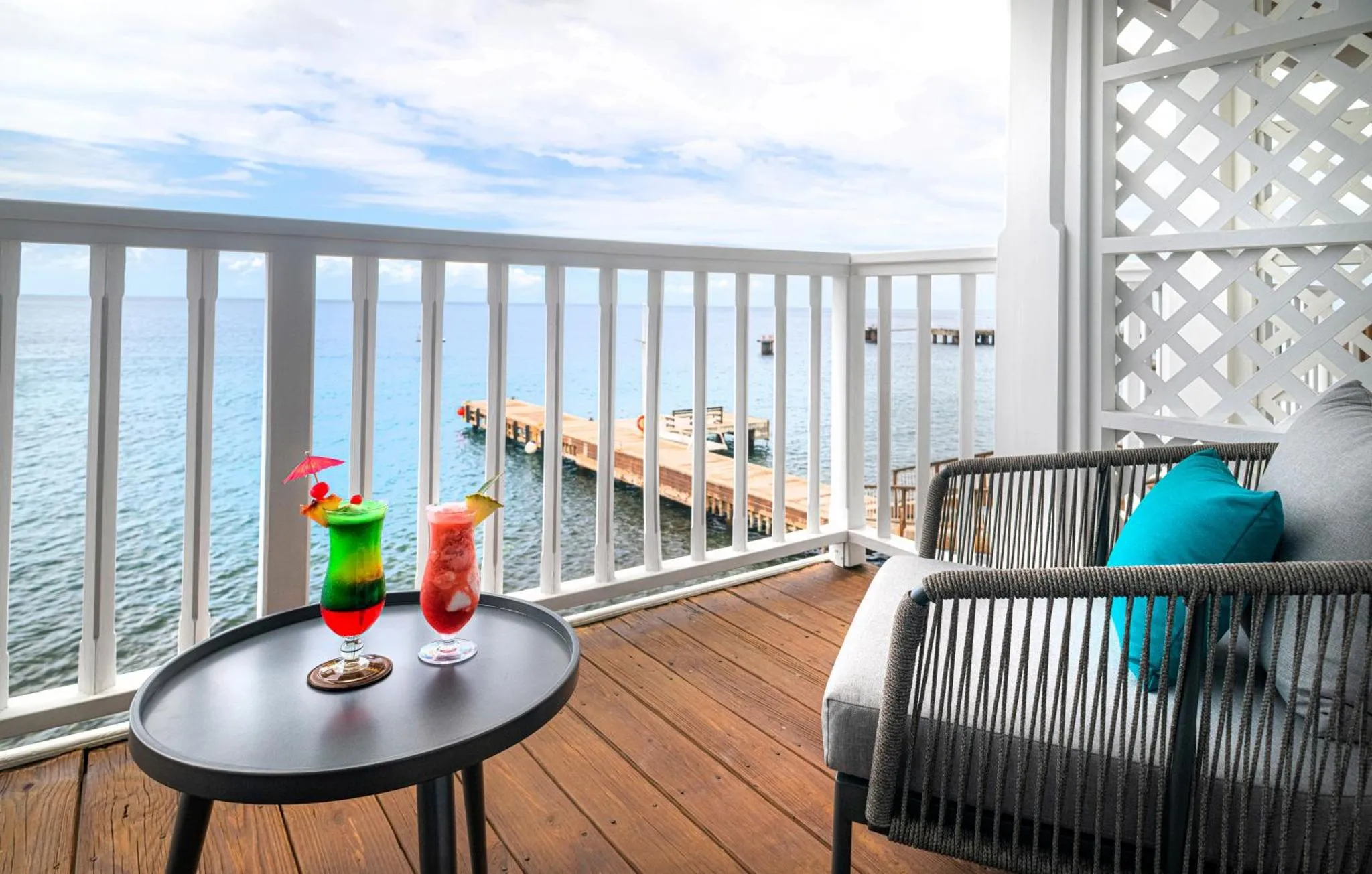 Une terrasse en bois avec des fauteuils et une table basse sur laquelle reposent des cocktails colorés, donnant sur la mer.