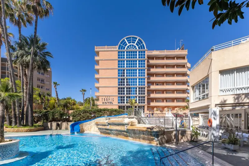 Vue d'un bâtiment beige avec des palmiers et une piscine au premier plan.