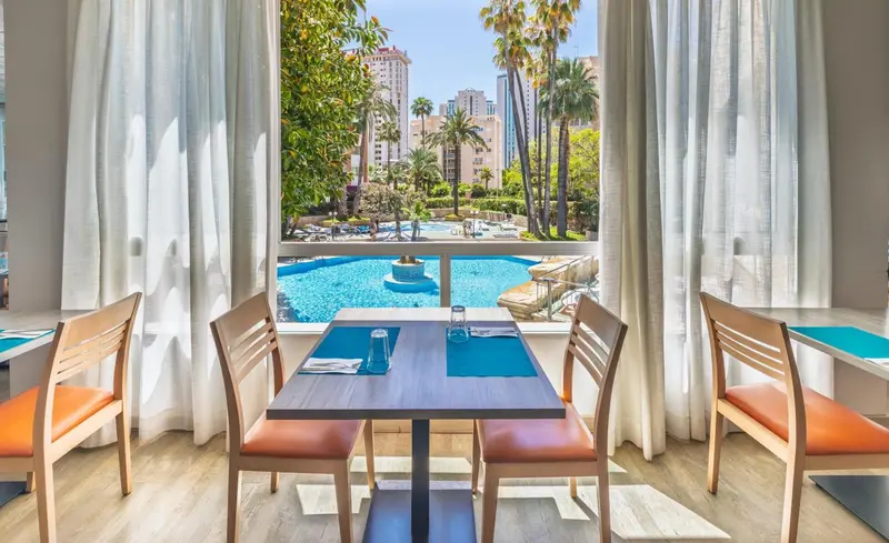 Une table est dressée devant une grande baie vitrée donnant sur une piscine et une végétation luxuriante.