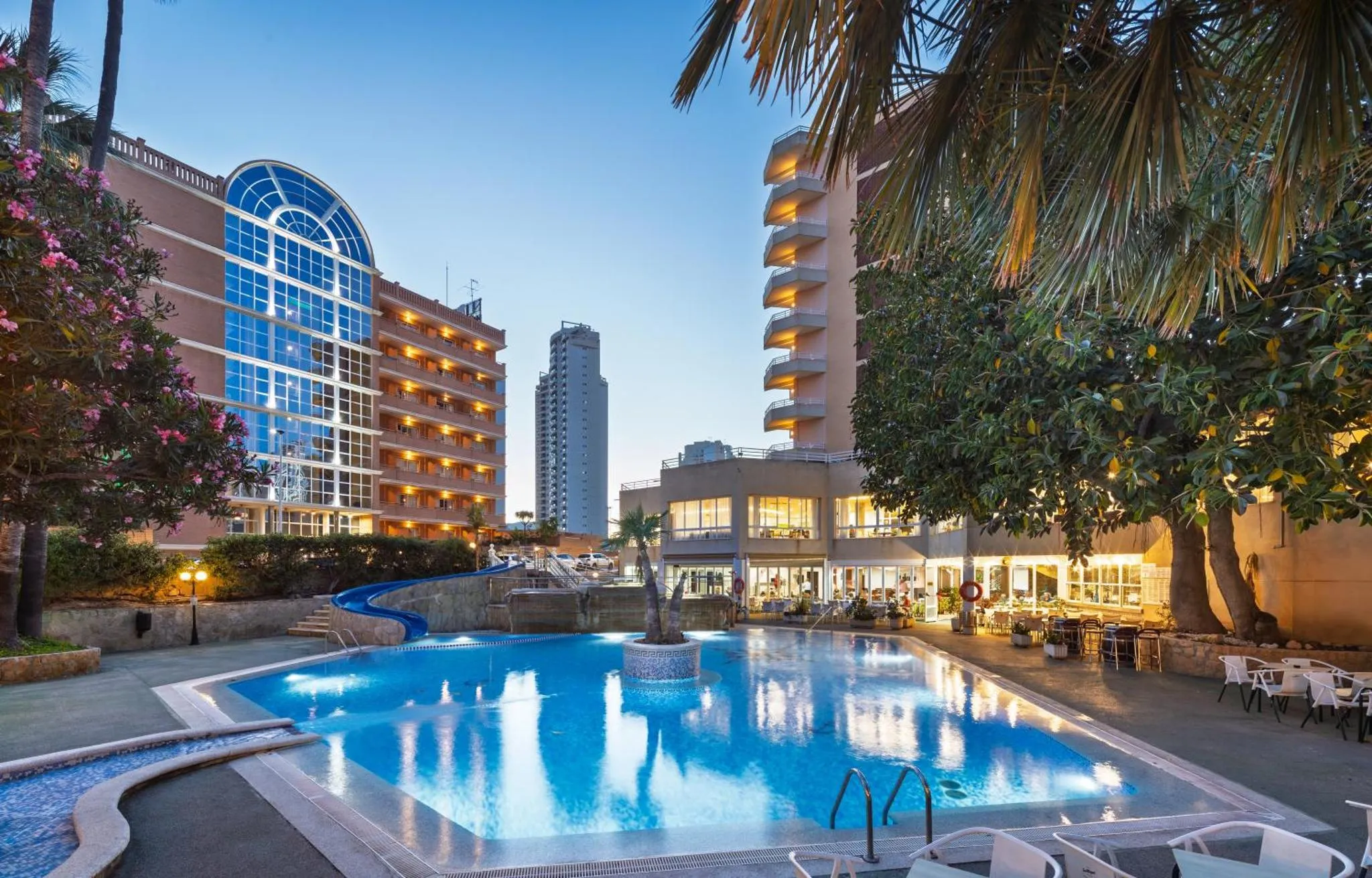 Bâtiment illuminé avec piscine et végétation luxuriante au crépuscule.