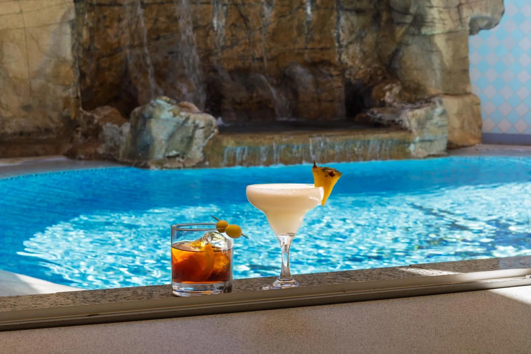 Deux cocktails colorés sont posés au bord d'une piscine turquoise avec une cascade rocheuse en arrière-plan.