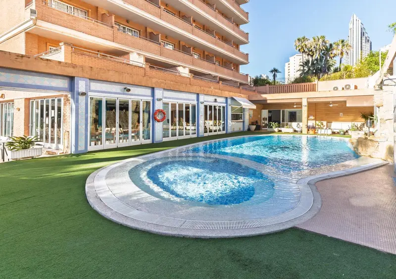 Piscine turquoise et terrasse ensoleillée devant un bâtiment résidentiel.