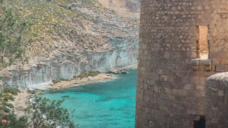 Vue d'une crique turquoise encadrée par des falaises rocheuses et une tour en pierre.