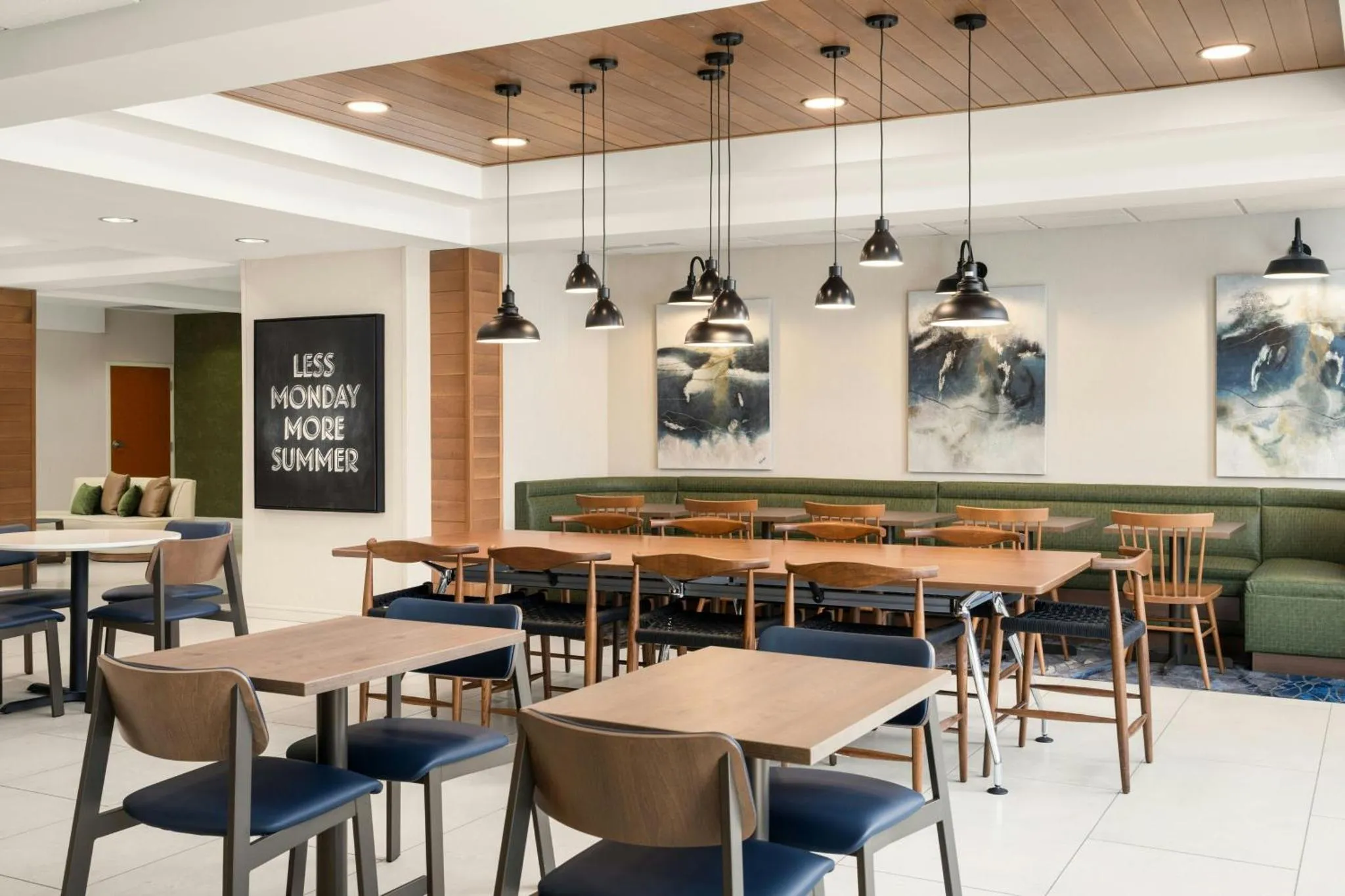 Une salle lumineuse avec des tables et des chaises en bois, des suspensions noires et des banquettes vertes.