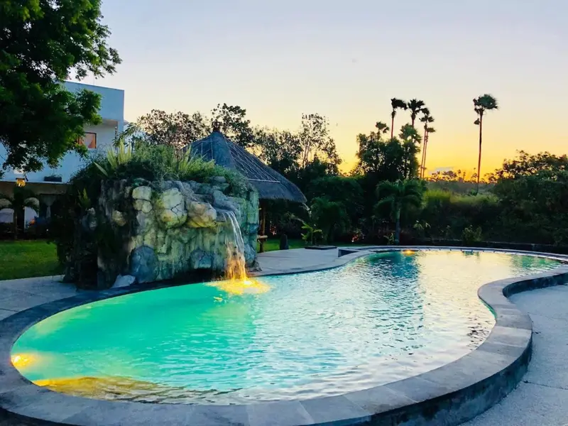 Une piscine avec une cascade artificielle et une végétation luxuriante au coucher du soleil.