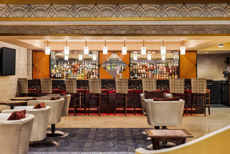 Un long comptoir de bar est aligné de bouteilles et de tabourets, avec des fauteuils et des tables autour.