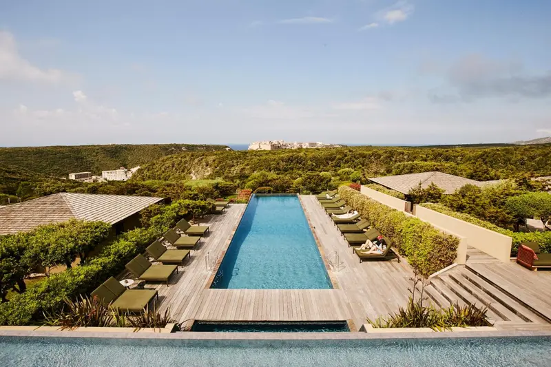 Une piscine rectangulaire bordée de transats offre une vue sur la végétation et la mer.