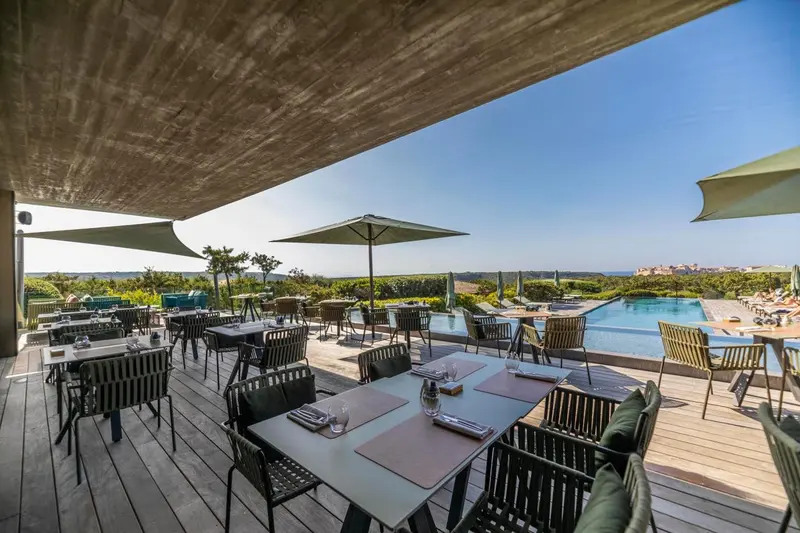 Terrasse ombragée avec tables dressées face à une piscine et un paysage côtier.