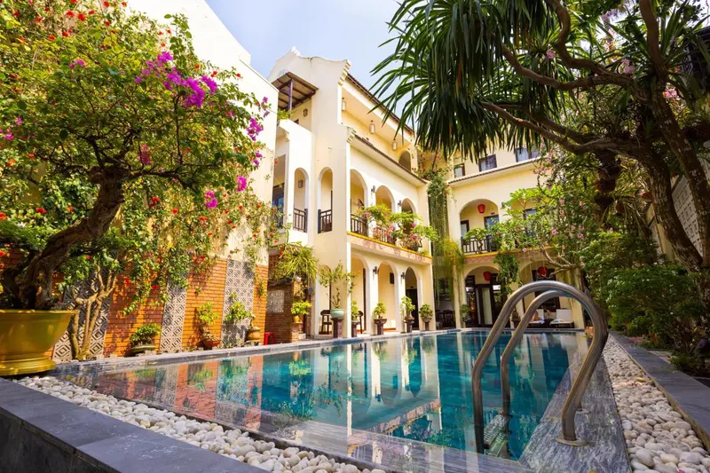 Une piscine turquoise bordée d'une terrasse ensoleillée devant un bâtiment blanc orné de végétation.