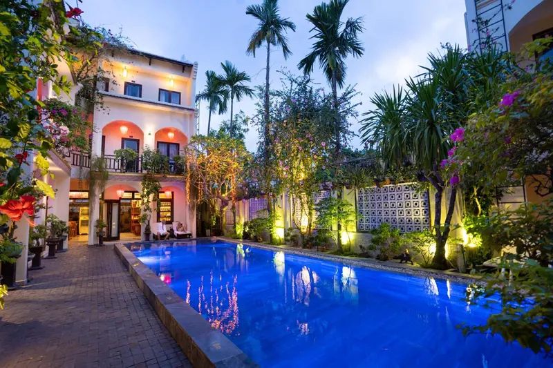 Une piscine turquoise est entourée d'une végétation luxuriante et d'un bâtiment éclairé.