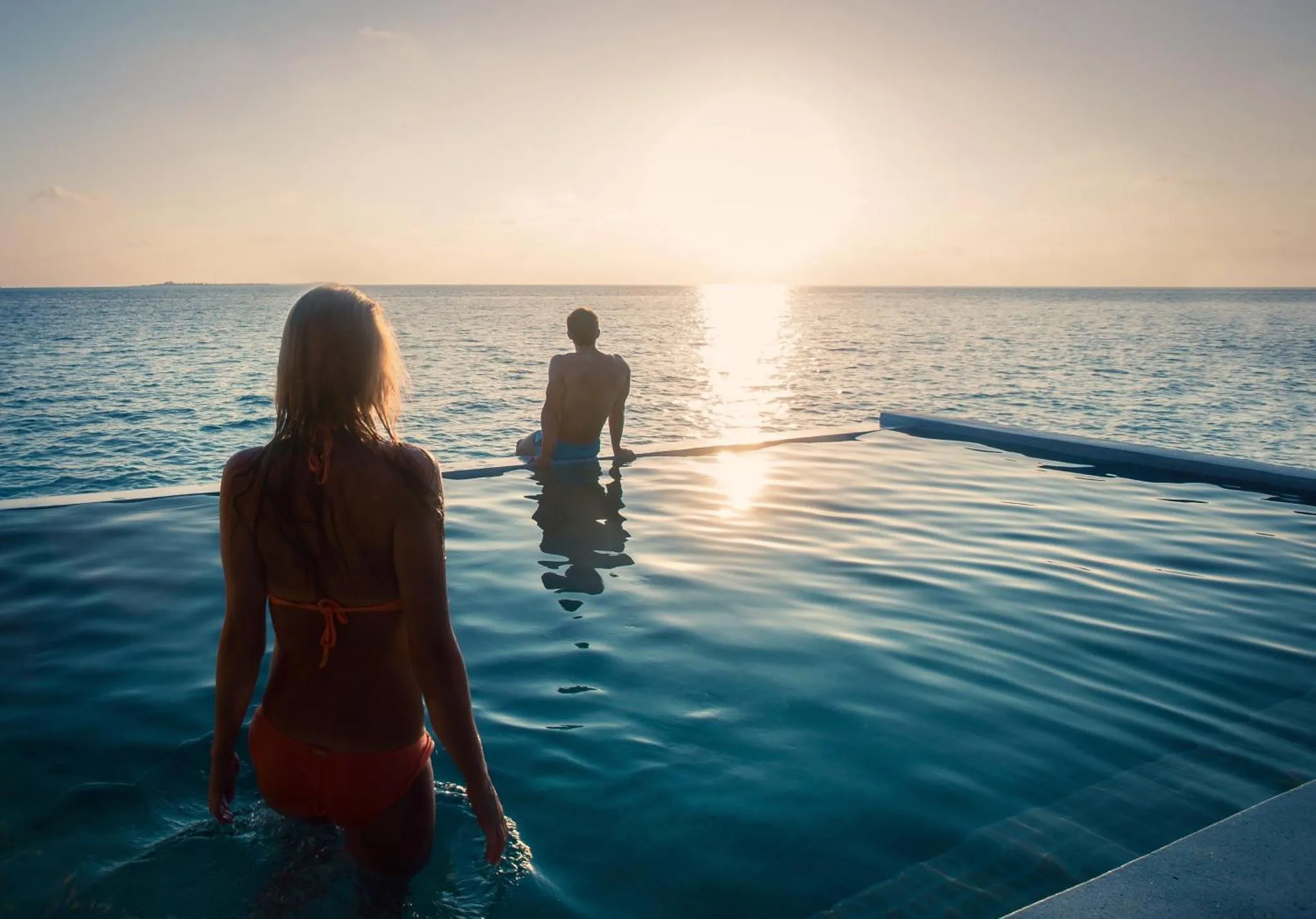 Deux personnes se tiennent au bord d'une piscine à débordement face à la mer au coucher du soleil.