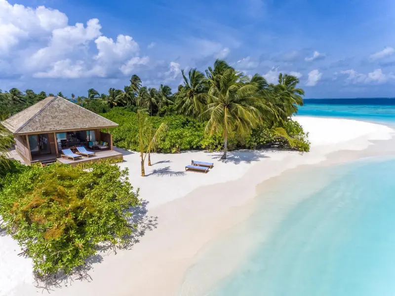 Vue aérienne d'une plage de sable blanc bordée de végétation tropicale et d'une villa sur pilotis.