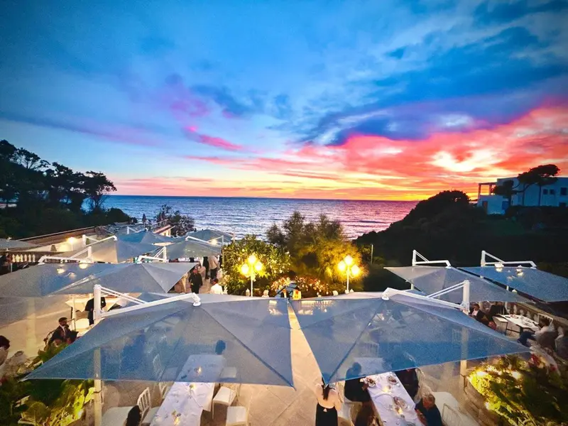 Terrasse ombragée avec tables dressées face à la mer au coucher du soleil.