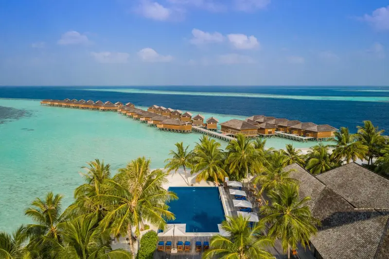 Vue aérienne d'un complexe hôtelier avec des villas sur pilotis et une piscine turquoise.