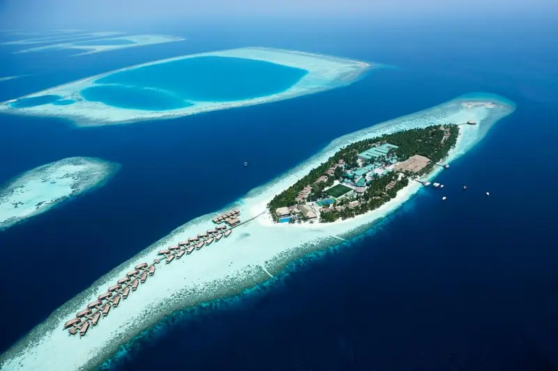 Vue aérienne d'une île tropicale avec des bungalows sur pilotis et une eau turquoise.