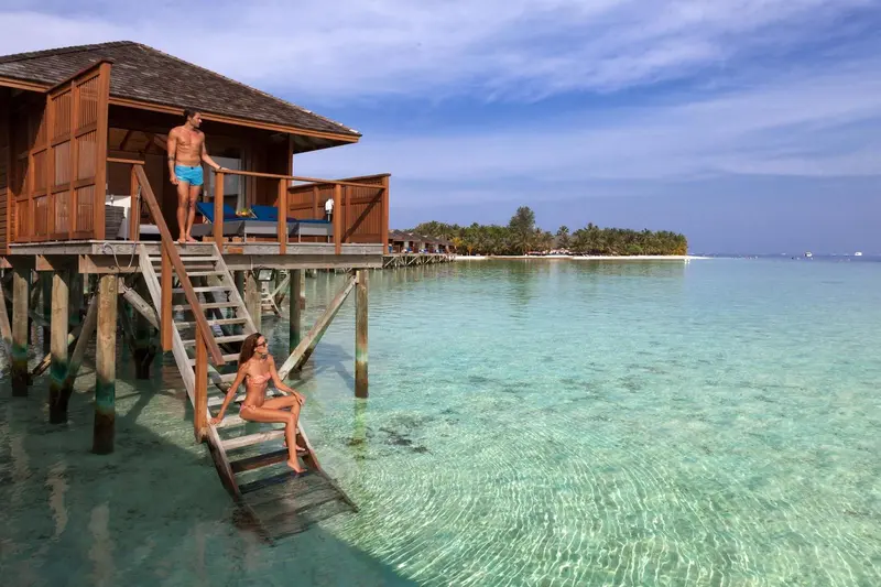 Deux personnes se tiennent sur la terrasse d'une maison sur pilotis au-dessus d'une eau turquoise.