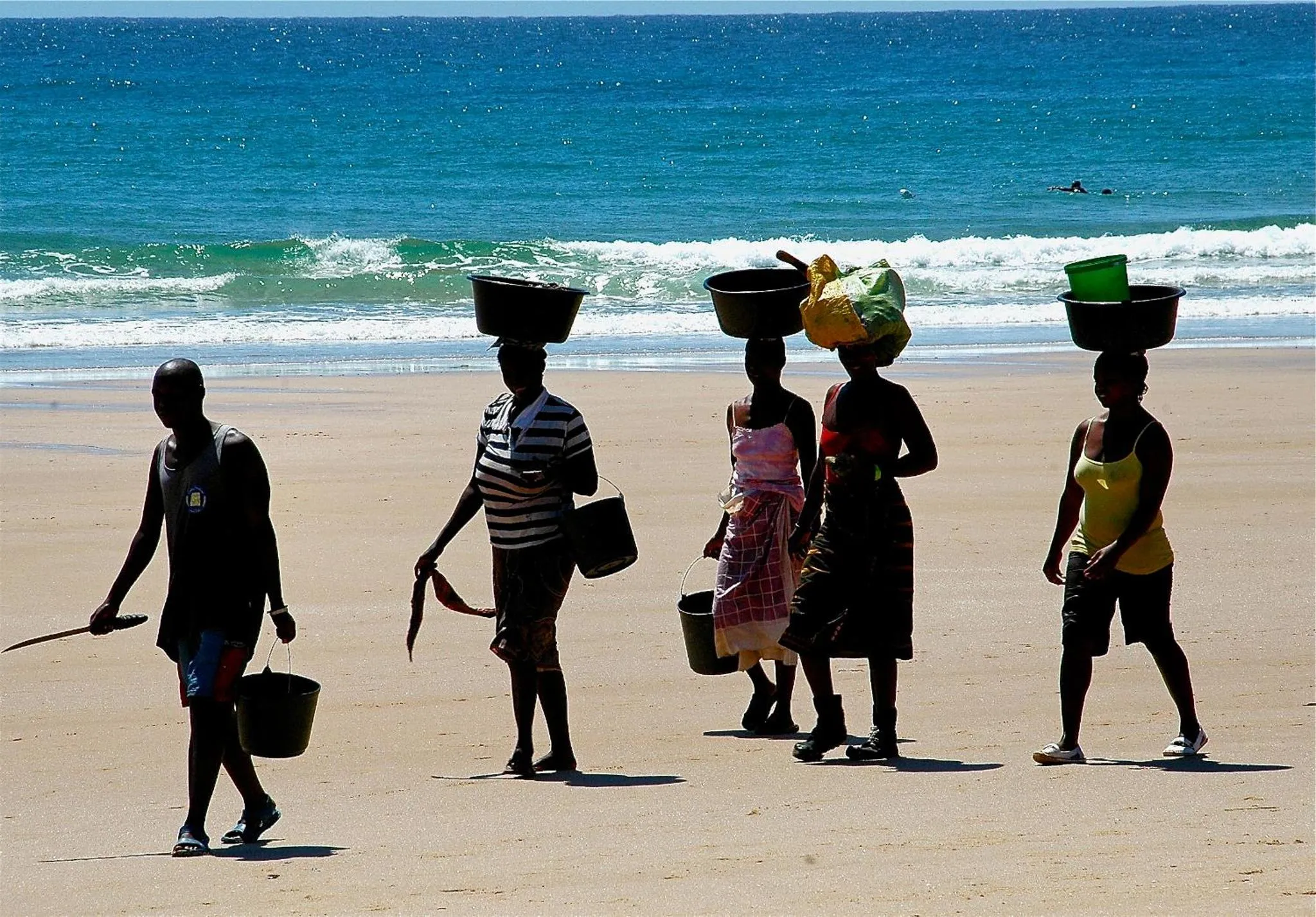 Une rangée de personnes marchant sur une plage de sable avec des paniers sur la tête.