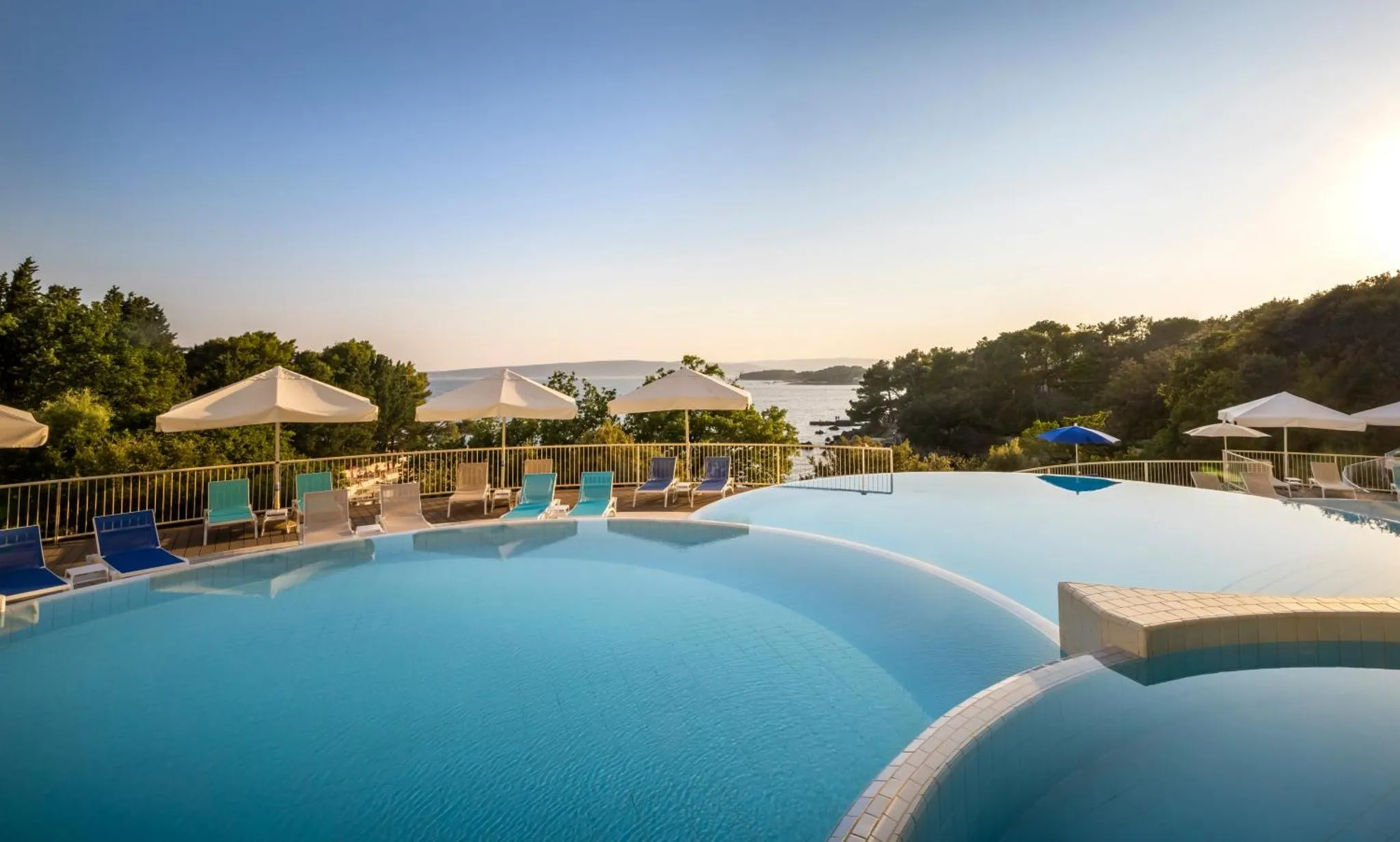 Piscine à débordement avec transats et parasols, vue sur la mer au loin.
