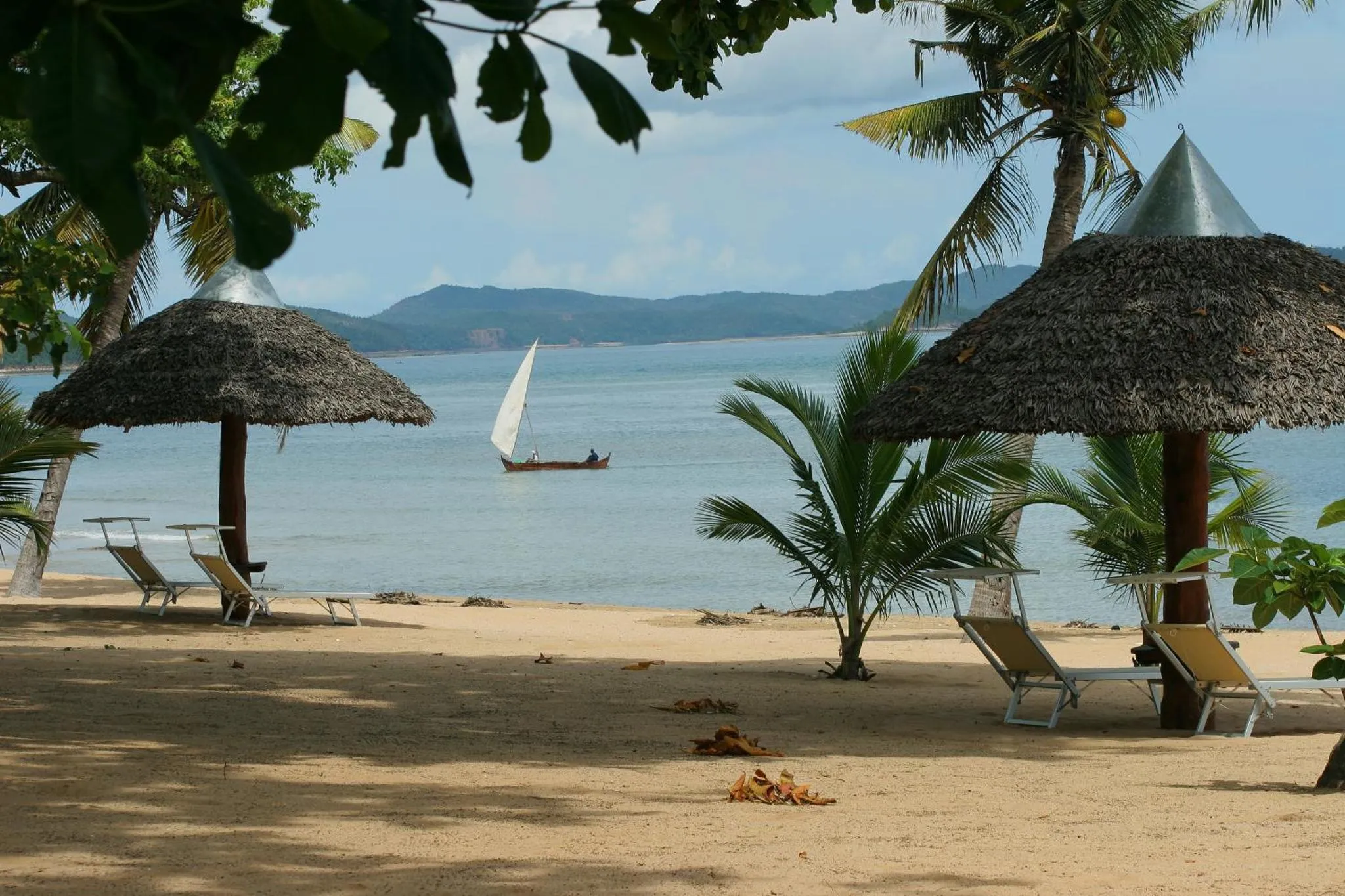 Plage de sable fin avec transats et parasols en chaume, vue sur la mer et un voilier.