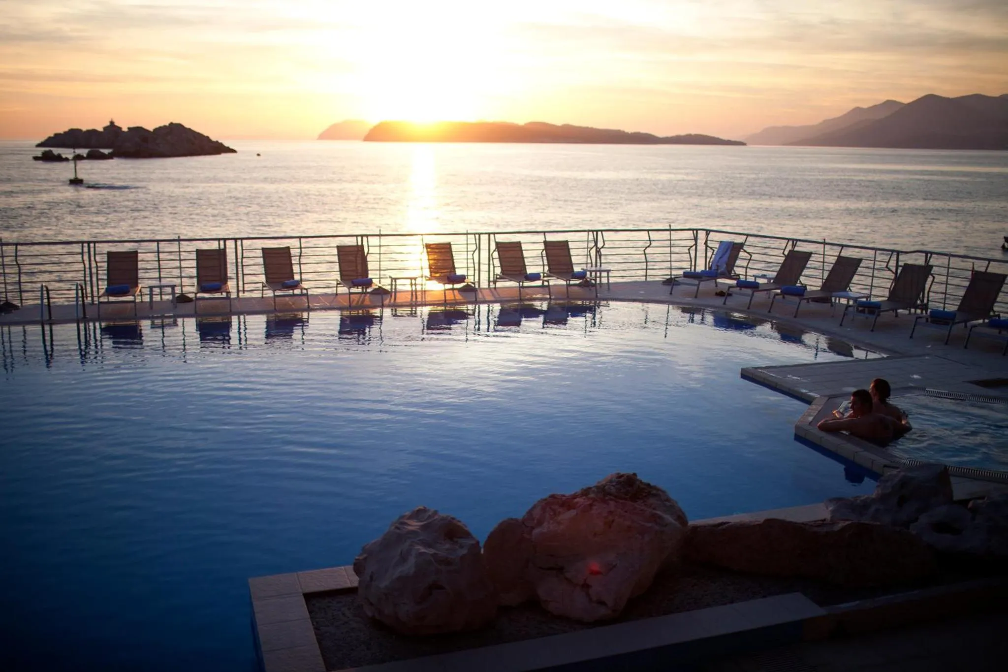 Transats bleus alignés au bord d'une piscine à débordement face à la mer au coucher du soleil.