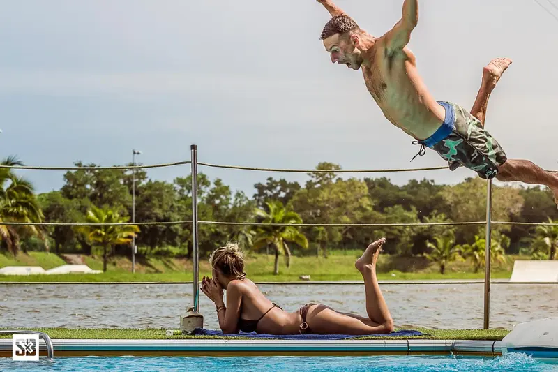 Une personne saute d'une plateforme vers une piscine, tandis qu'une autre se détend au bord.