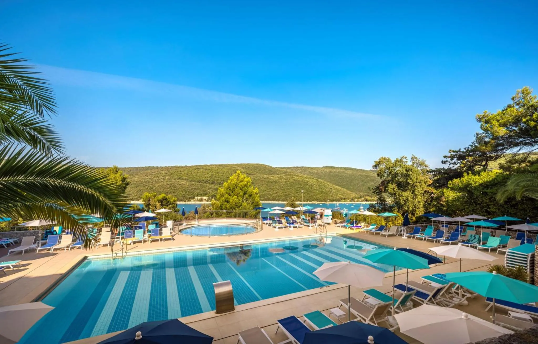 Une piscine turquoise bordée de transats sous des parasols bleus, avec une vue sur la mer et une colline verdoyante.