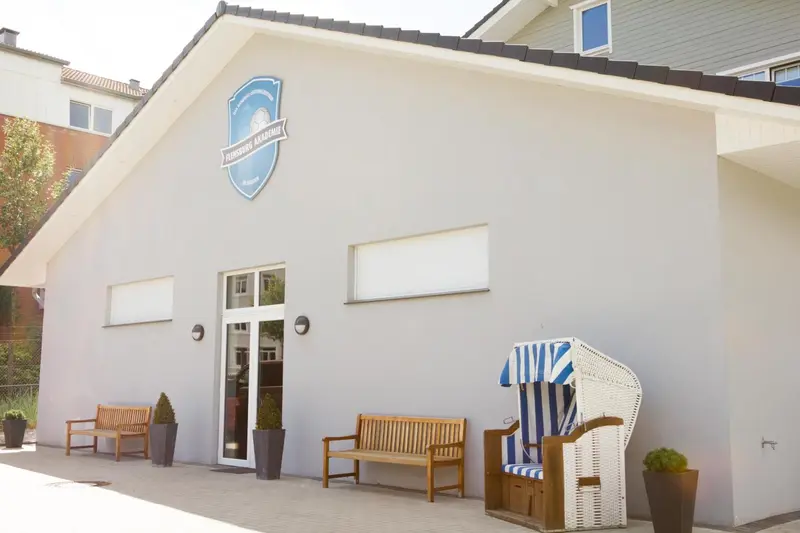 Façade blanche d'un bâtiment avec des bancs et un fauteuil en osier sur une terrasse.