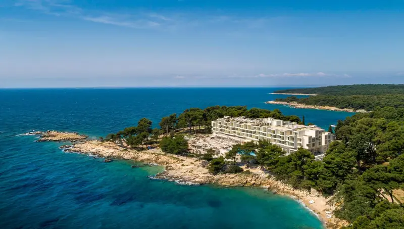 Vue aérienne d'un bâtiment blanc au bord de la mer turquoise et d'une côte rocheuse verdoyante.