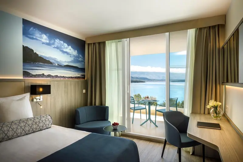 Chambre lumineuse avec un lit fait et une vue sur la mer depuis une baie vitrée.