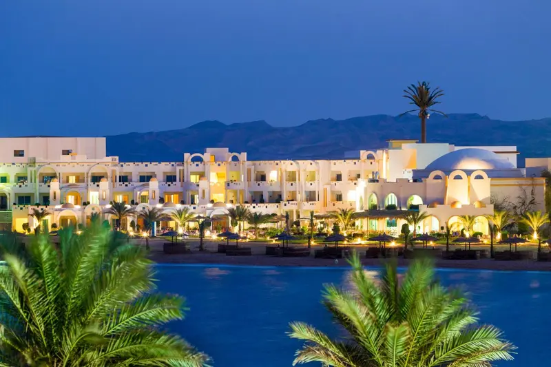 Bâtiment blanc illuminé au bord de l'eau, avec végétation tropicale au premier plan et montagnes en arrière-plan.