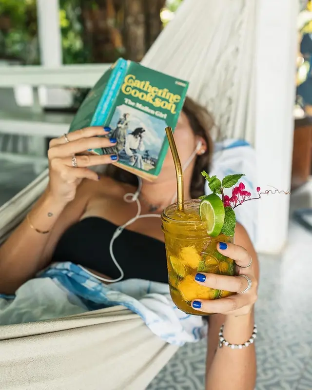 Une femme se détend dans un hamac avec un livre et une boisson.